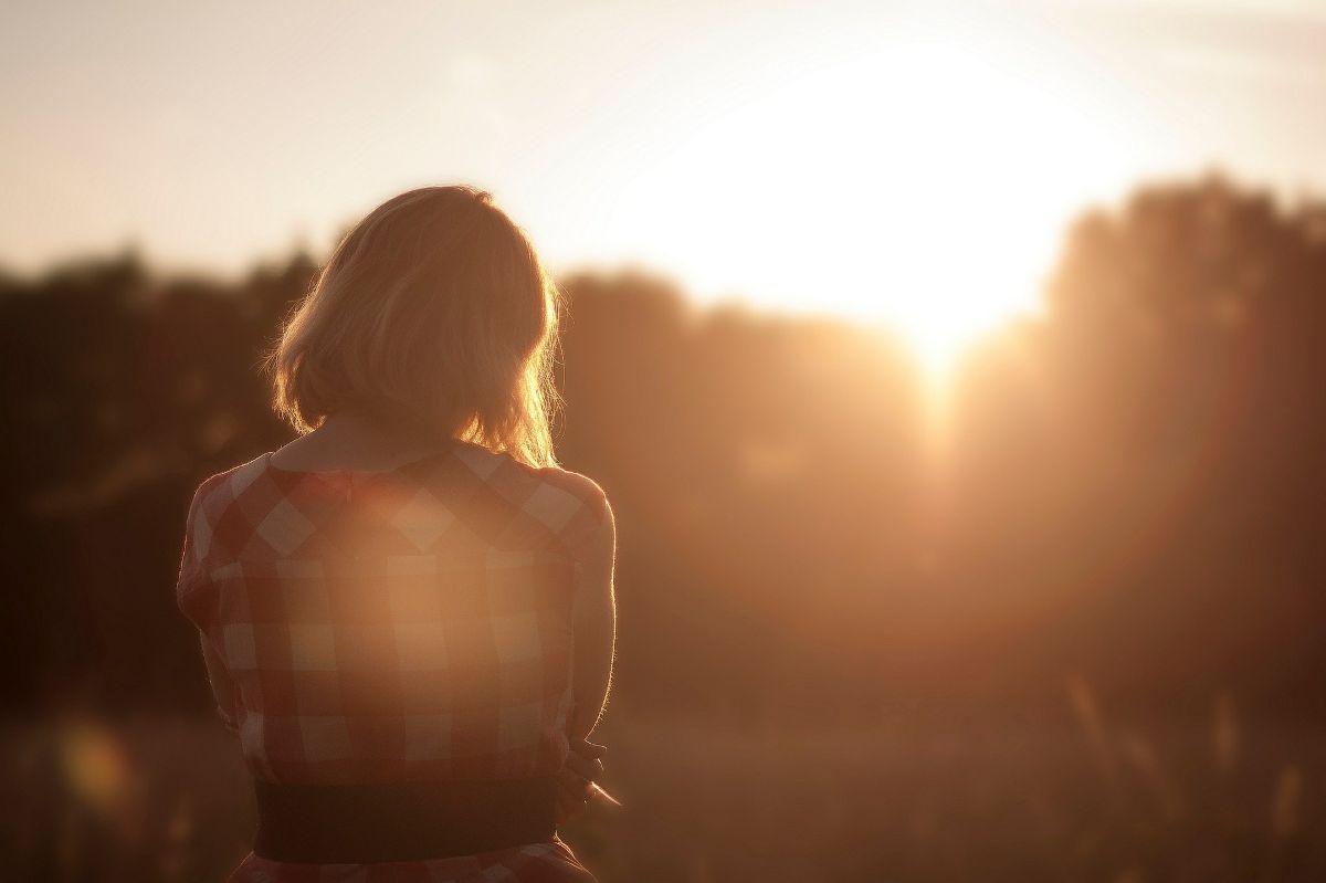 une femme debout de dos au couché du soleil tête basse et mains repliées sur elle